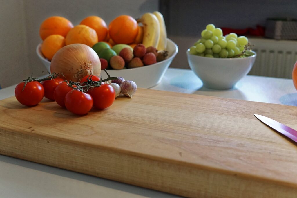 A wooden cutting board with tomatoes, garlic, and a knife, with bowls of fruit in the background.