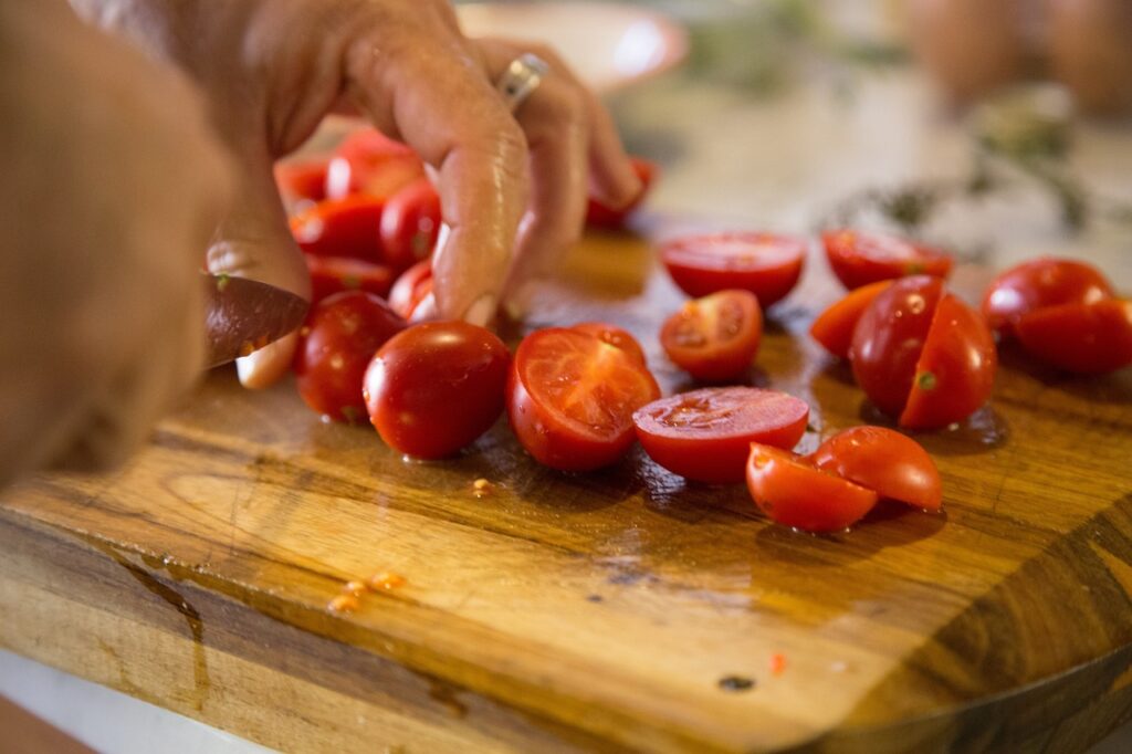 Hands slicing cherry tomatoes on a wooden cutting board.