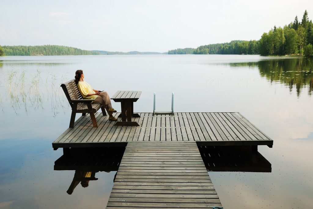 Person sitting on a bench by a calm lake with a wooded shoreline in the distance.